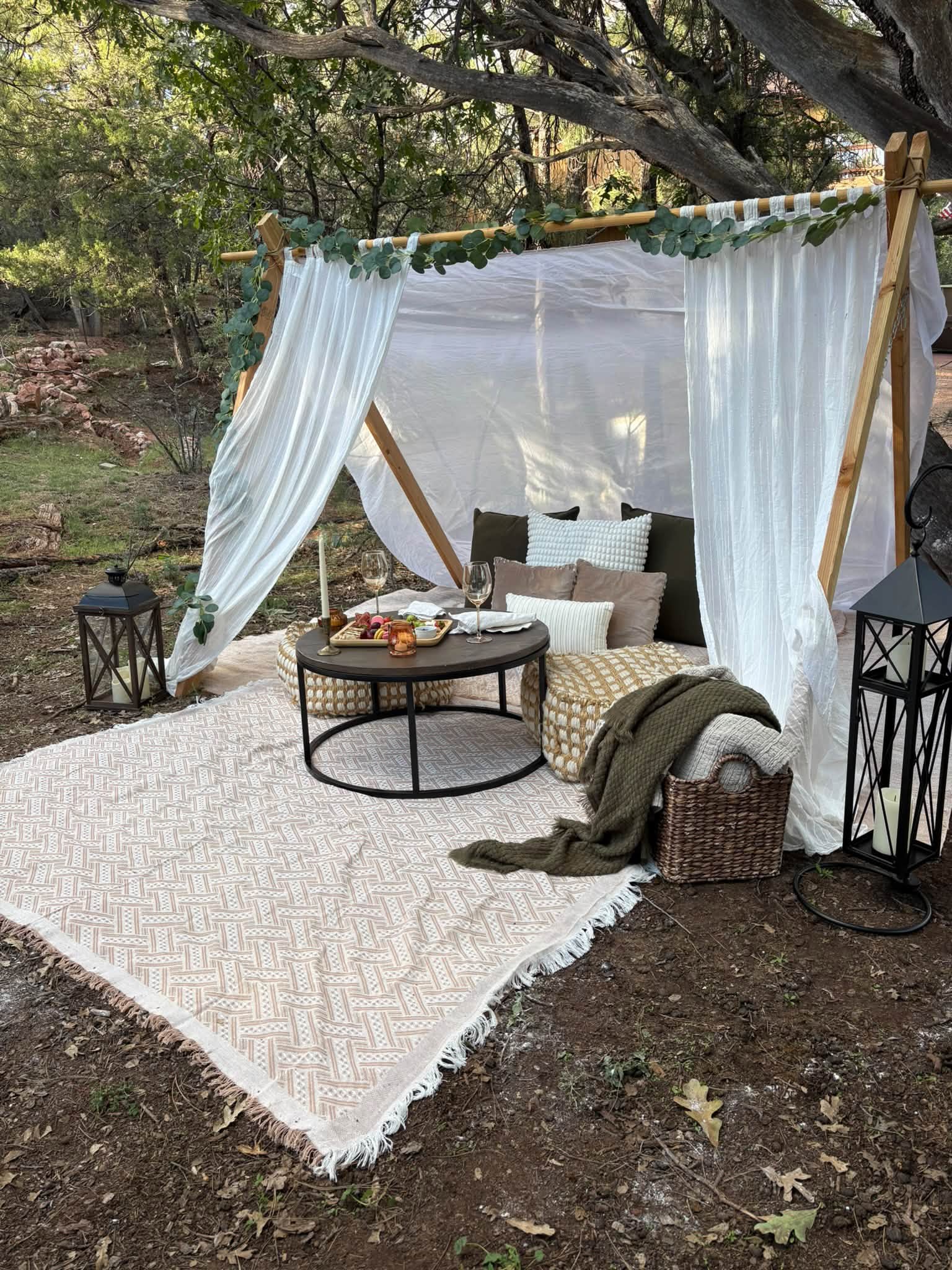 An intimate styled setting with linen curtains, lanterns, cushions and a low table in the woods